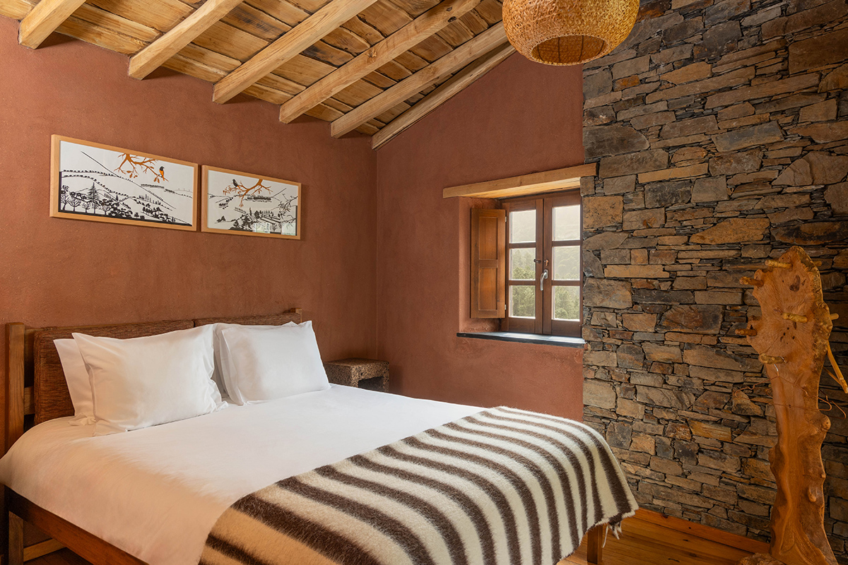 A schist-stone cottage bedroom in Cerdeira: terracotta walls, exposed wood beams, white linens, a striped wool throw at the foot of the bed, and framed bird artwork.