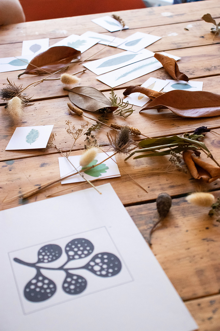 A wooden table at the studio: pressed leaves, dried botanicals, color swatches in soft seafoam, and a finished hand-printed botanical artwork in dark indigo on cream paper.